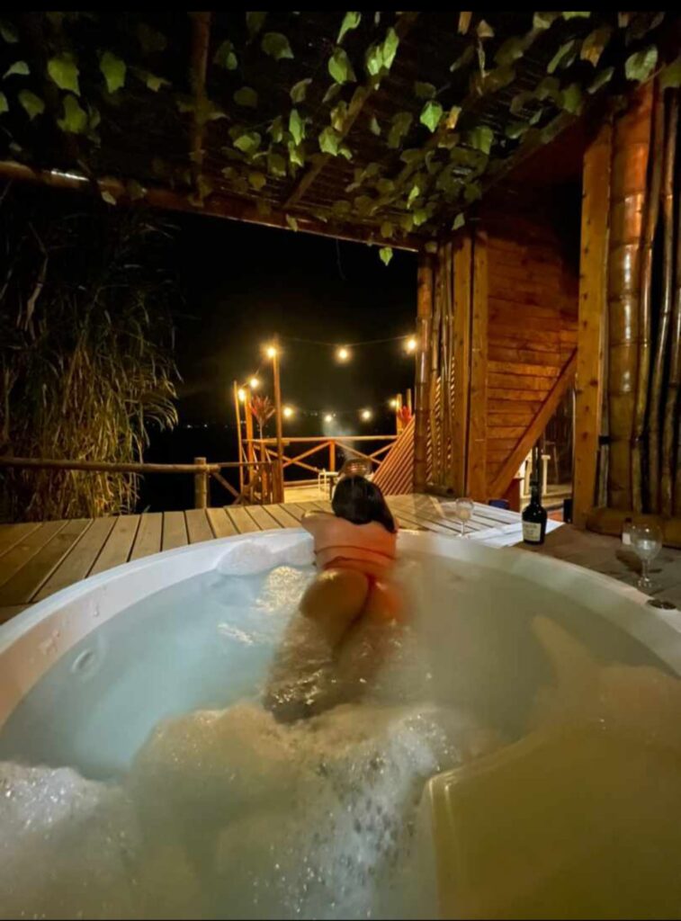 A woman lounges in a bubbling jacuzzi at night, facing out toward the dark mountain landscape in Colombia, under warm string lights and a rustic wooden canopy
