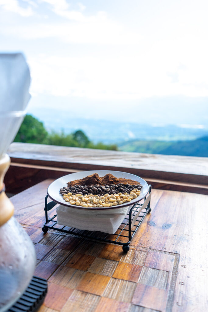 A plate displaying different stages of Colombian coffee beans—raw, roasted, and ground—set on a wooden table at a coffee shop, with a scenic view of the Colombian mountains and sky in the background