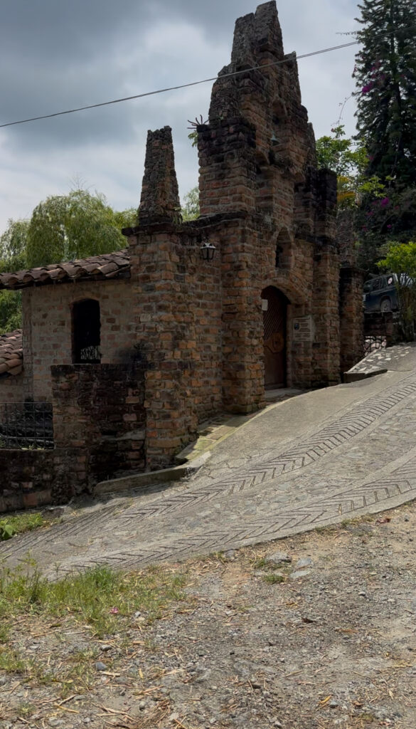 A rustic stone church built into the mountainside in Colombia, with tall, weathered brick towers and a steep, patterned stone path leading up to its wooden door under a cloudy sky